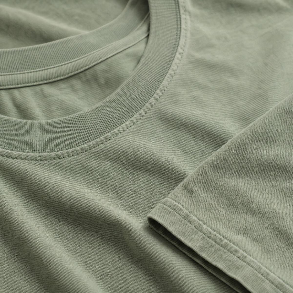 Close-up of a folded sage green t-shirt showing the ribbed collar, hem and garment-dyed cotton weave